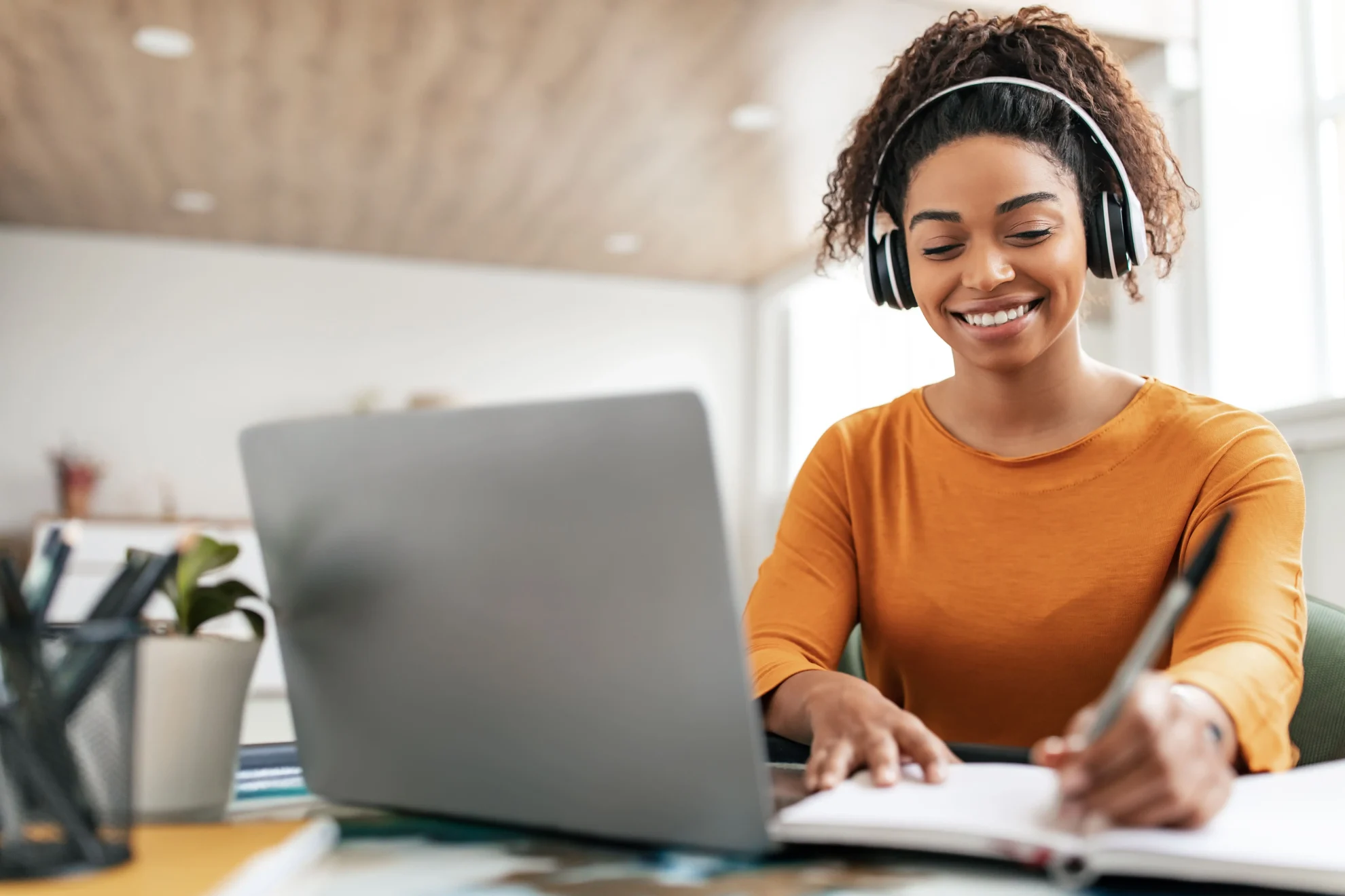 Mulher sorrindo sentada em frente a um computador, usando headphone e fazendo anotações em um caderno. Ela está sorrindo.