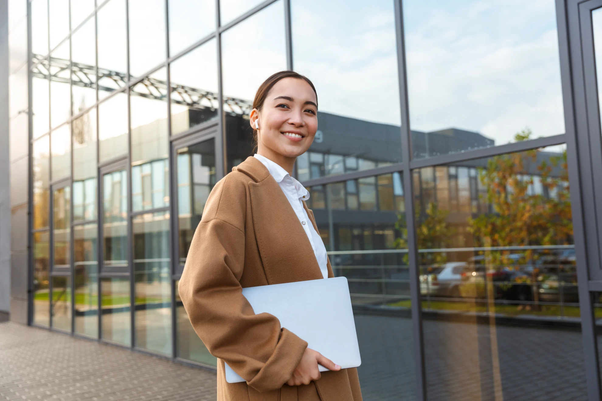 Mulher em frente a um prédio empresarial sorrindo e carregando um notebook.